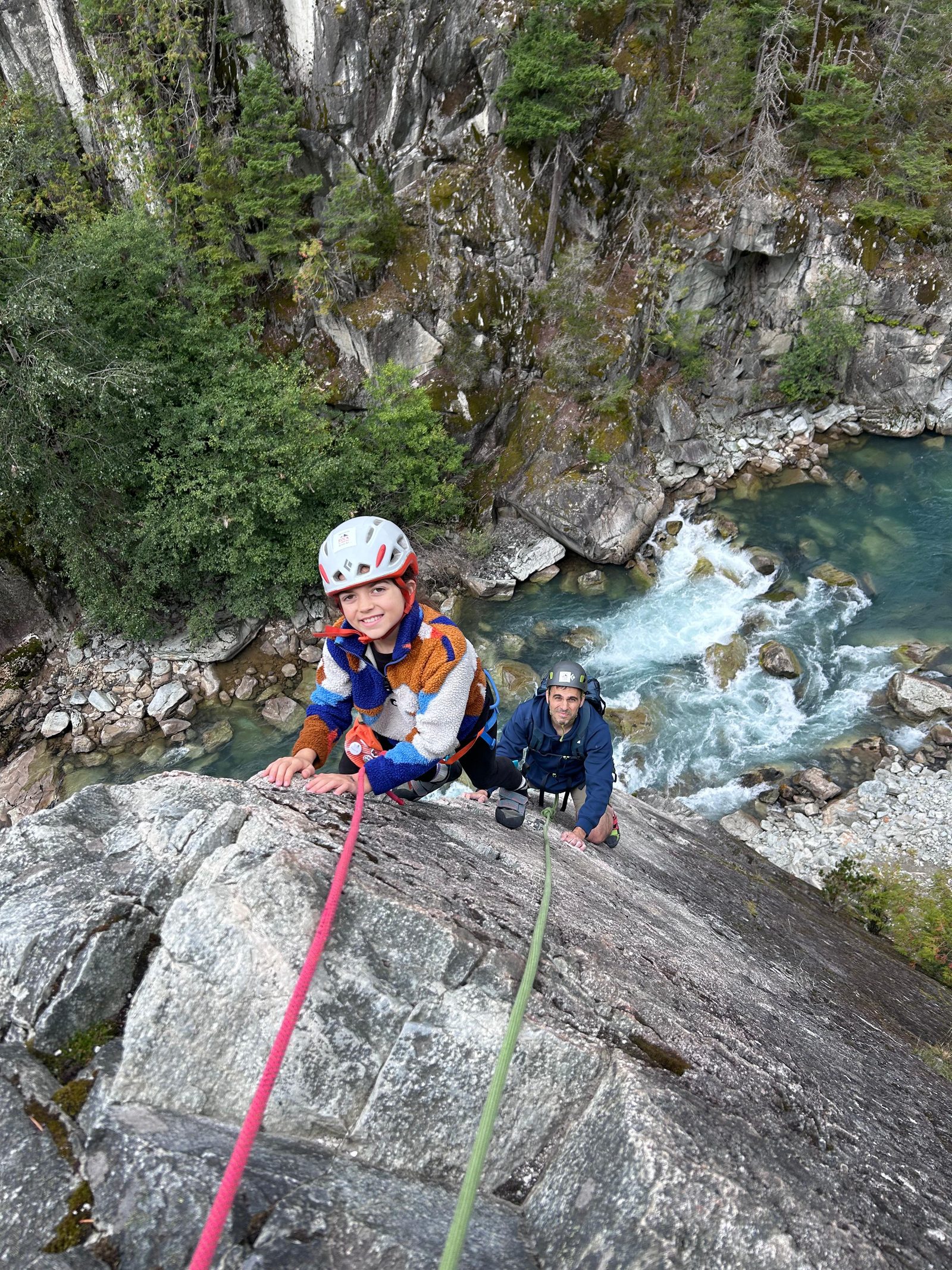 Alice in a white helmet and striped fleece, and Keith in a blue jacket and dark helmet, on a granite slab above a river in Squamish. A pink rope and a green rope run down the rock.