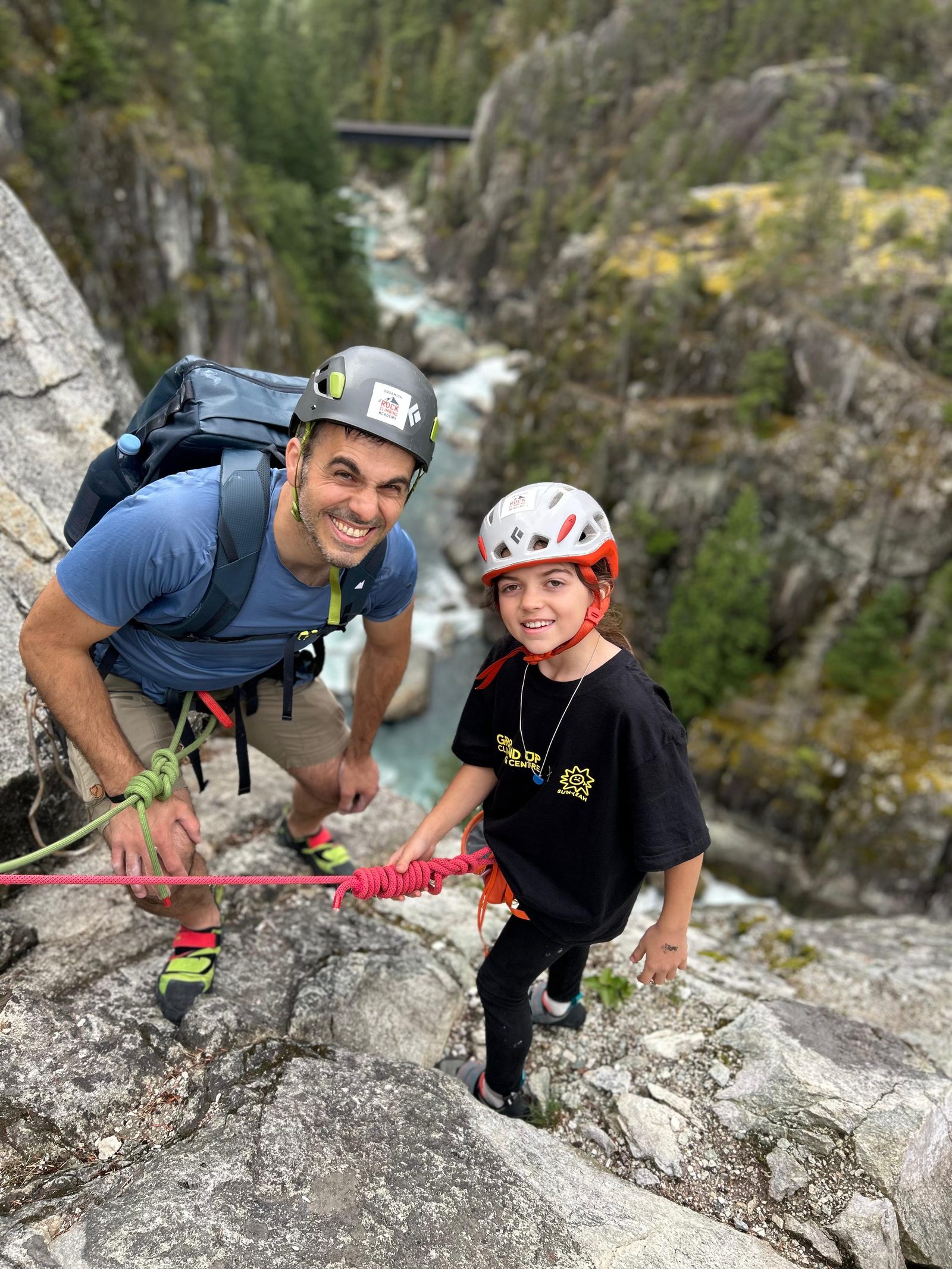 Keith and Alice on the boulders in Squamish, both in helmets and holding the ropes, with the river in the gorge behind them.