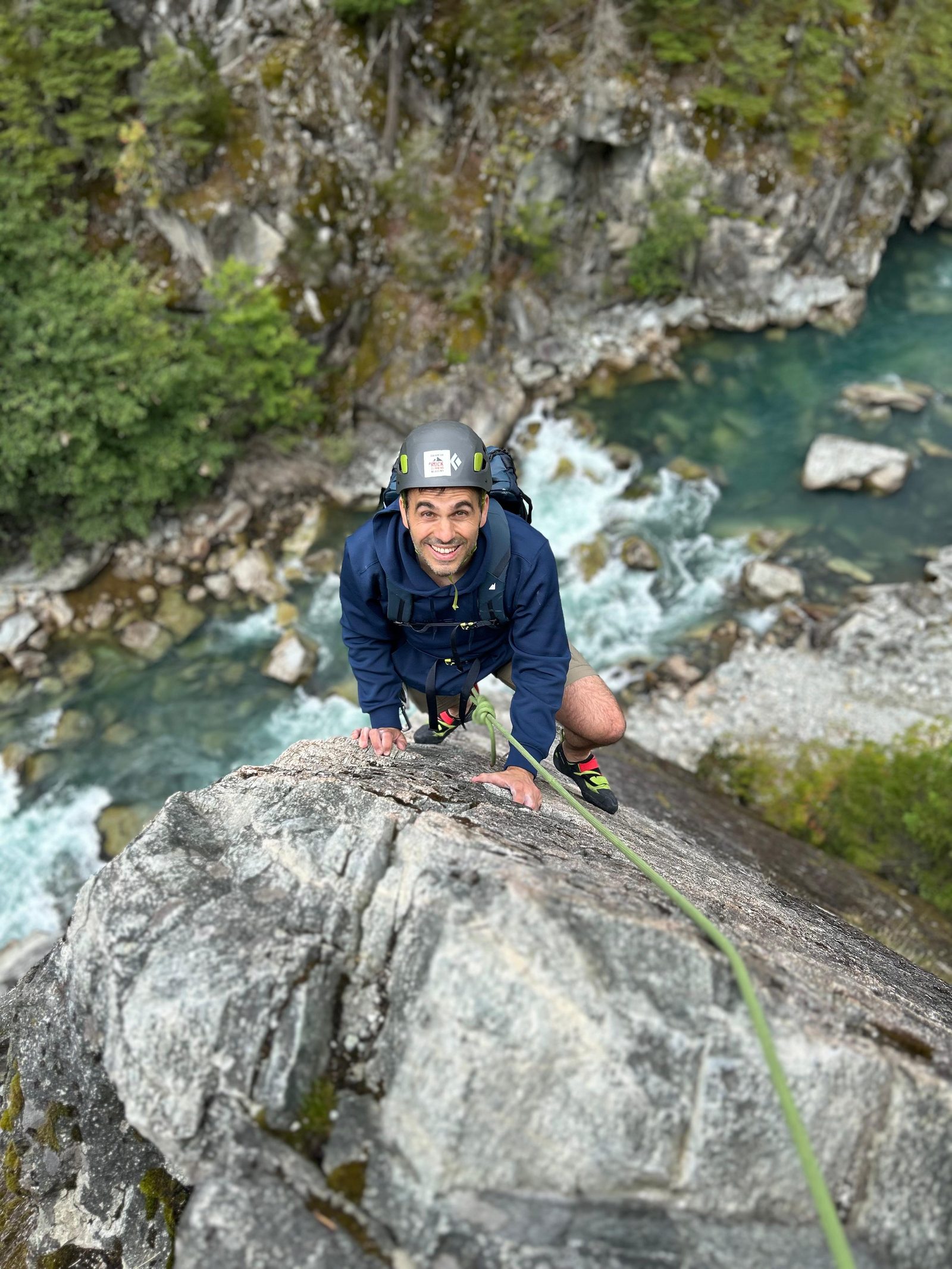 Keith at Star Chek, in a navy jacket and grey helmet, on a granite slab above the river in Squamish, with a green rope running down the rock.